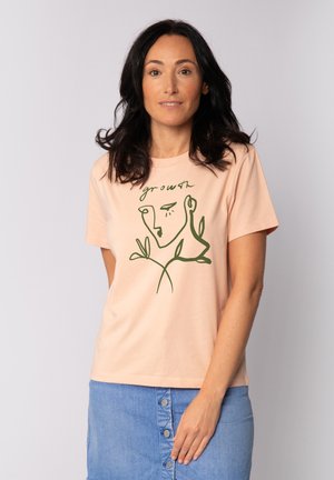 Perzikkleurige T-shirt met korte mouwen, gemaakt van zachte katoen, met een groene lijntekening van een gezicht omringd door planten en het woord "groei."
