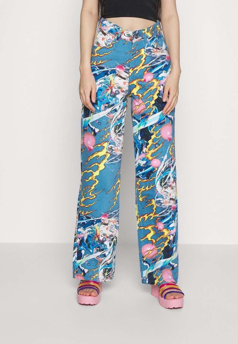 Džíny s širokými nohavicemi v denimové barvě, s modrou základnou a živými, pestrými abstraktními vzory s motivy blesku. Růžové platformové sandály.