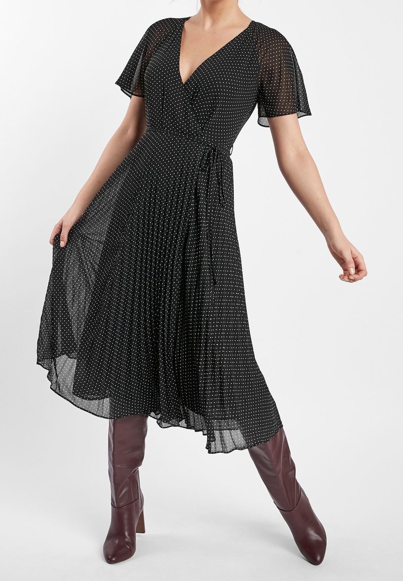 Robe portefeuille noire avec manches courtes évasées, pois blancs, texture plissée et ceinture à nouer à la taille, associée à des bottes bordeaux montant jusqu'au genou.