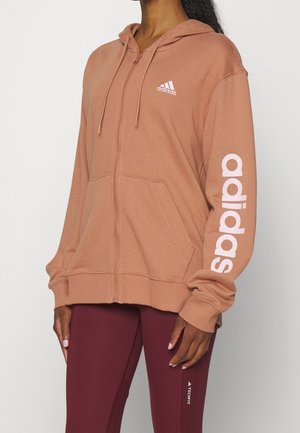 Persoon draagt een stoffig roze Adidas zip-up hoodie met logo op borst en mouw, gecombineerd met bordeauxrode Adidas Techfit leggings.