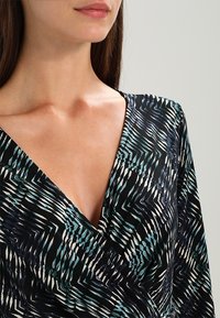 Damernas omslagstopp i svart tyg med turkosa och vita zigzag-mönster, V-ringning och mjuk, lätt textur.