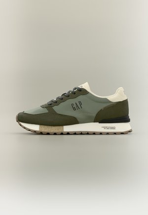 Zapatilla baja de color verde oliva y crema con cordones negros, suela de goma y la marca "GAP" en el lateral, diseñada para uso casual.