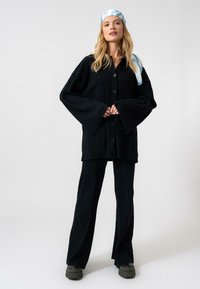 Zwarte geribde cardigan met klokmouwen, knoopsluiting, gecombineerd met bijpassende zwarte geribde broek. Een lichtblauwe met patronen hoofddoek maakt de look compleet.