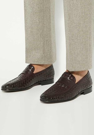 Brown woven leather loafers with a low heel, featuring a smooth toe cap and subtle stitching details. Combined with light gray trousers.