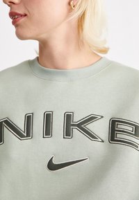 Ljusgrön sweatshirt med "NIKE"-logotyp i mörkgrön brodering och en liten Nike-swoosh. Har en ribbad rund halsringning.