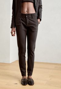 Pantaloni marroni su misura con una vestibilità slim, dotati di due bottoni frontali, tasche laterali e una texture liscia. Indossati con mocassini neri.