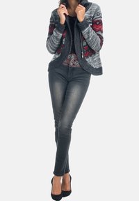 Šedý pletený kardigan s červenými a černými geometrickými vzory, na zip, s žebrovanými manžetami, nošený přes tmavý top a v kombinaci s černými úzkými džínami a podpatky.