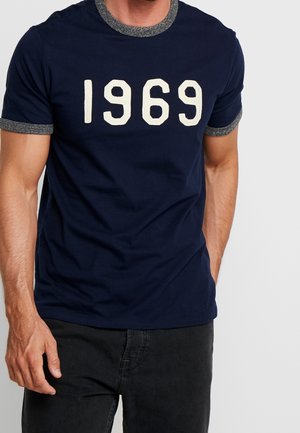 Mann trägt ein navyblaues T-Shirt mit grauem Kragen und Ärmelsäumen, das große weiße Zahlen "1969" auf der Brust hat.