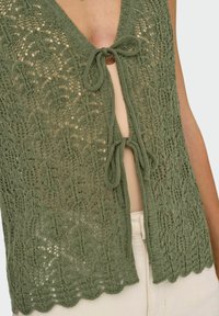 Oliwkowy dzianinowy top z delikatnym wzorem z koronki, dekoltem w kształcie litery V oraz zawiązywaniem na kokardkę. Falbaniaste krawędzie dodają tekstury do projektu.
