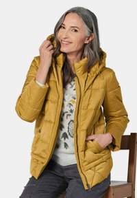 Gelbe Puffermantel mit Kapuze, ausgestattet mit einem gesteppten Muster, einem Reißverschluss auf der Vorderseite und seitlichen Taschen. Trägt man über einem leichten Pullover.