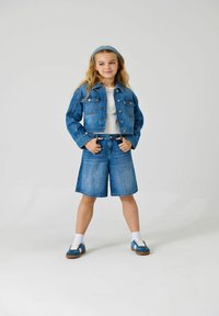Lichte blauwe denimjack en short set. Het jack heeft knoopsluitingen en twee voorzakken. Het model draagt een witte top en blauwe schoenen.