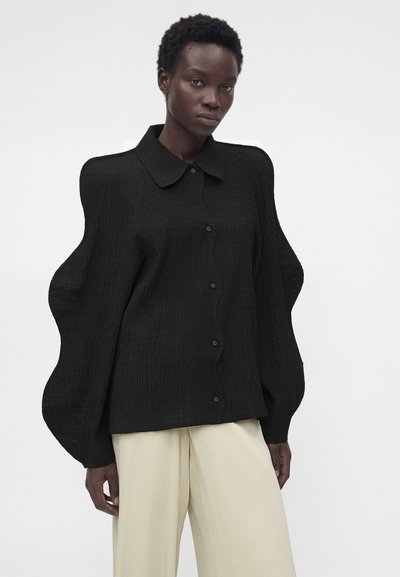 Γυναίκα με σκουρόχρωμο δέρμα που φοράει μαύρο υφασμάτινο φαρδύ μπλουζάκι με υπερβολικά μανίκια και μπεζ παντελόνι με φαρδιά μπατζάκια, στέκεται σε απλό φόντο.