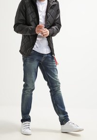 Svart quiltad jacka med huva, bärs över en vit prickig t-shirt, tillsammans med blå denimjeans och vita sneakers.