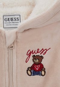 Chaqueta con cremallera de forro polar color rosa claro con un interior suave. Presenta un oso bordado con un suéter rojo, con "guess" cosido en rojo.