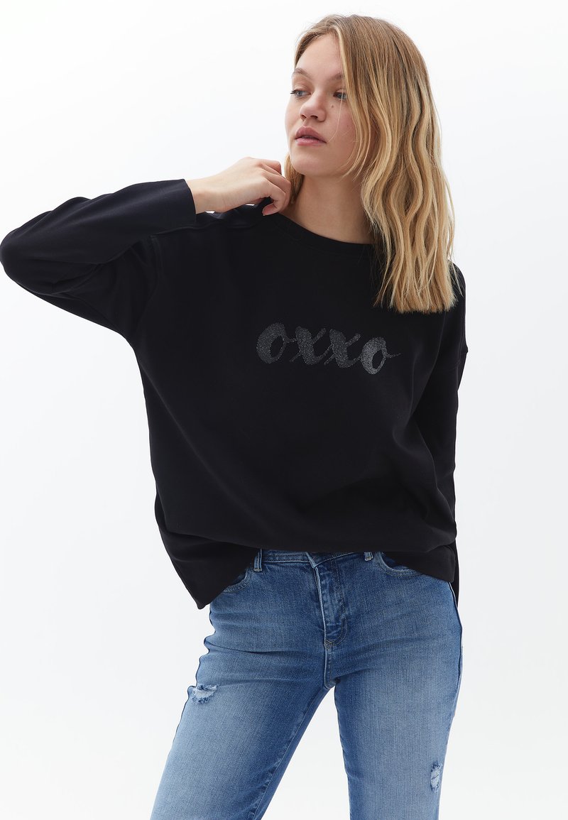 Czarny sweter z napisem "oxxo" w błyszczącej czcionce, w połączeniu z niebieskimi, zniszczonymi dżinsami. Charakteryzuje się luźnym krojem i długimi rękawami.