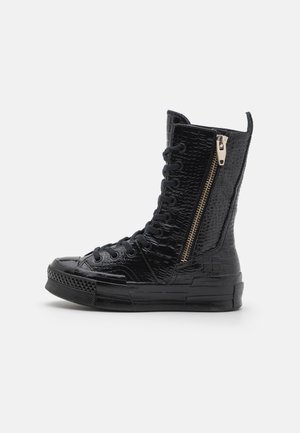 Bottine sneaker noire montante avec surface texturée, lacets à l’avant, semelle en caoutchouc et fermeture éclair latérale sur fond blanc.