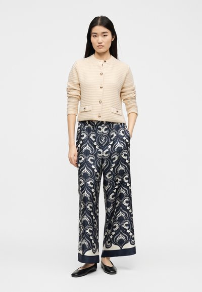 Γυναίκα όρθια, φοράει μπεζ πλεκτή ζακέτα με κουμπιά, μπλε σκούρο, φαρδιά παντελόνια με σχέδια και μαύρα παπούτσια με φλατ σόλα, μπροστά σε λευκό φόντο.