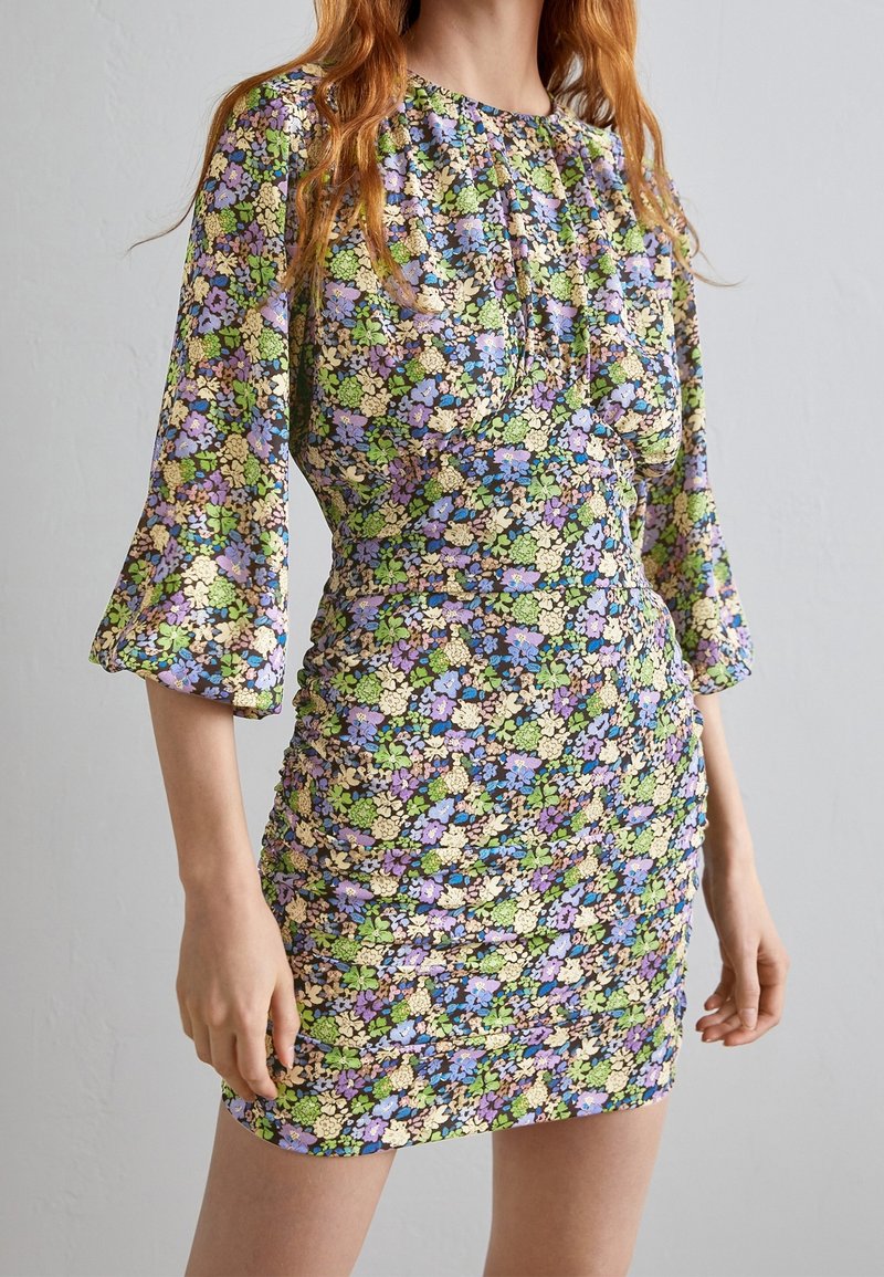 Femme portant une robe fleurie ajustée avec des fleurs pastel violettes, vertes et crème, des manches trois-quarts et des détails froncés à la taille.
