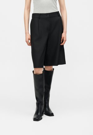 Kobieta w czarnych, dopasowanych szortach do kolan, jasnoszarym bezrękawniku oraz czarnych, skórzanych kozaczkach za kolano.