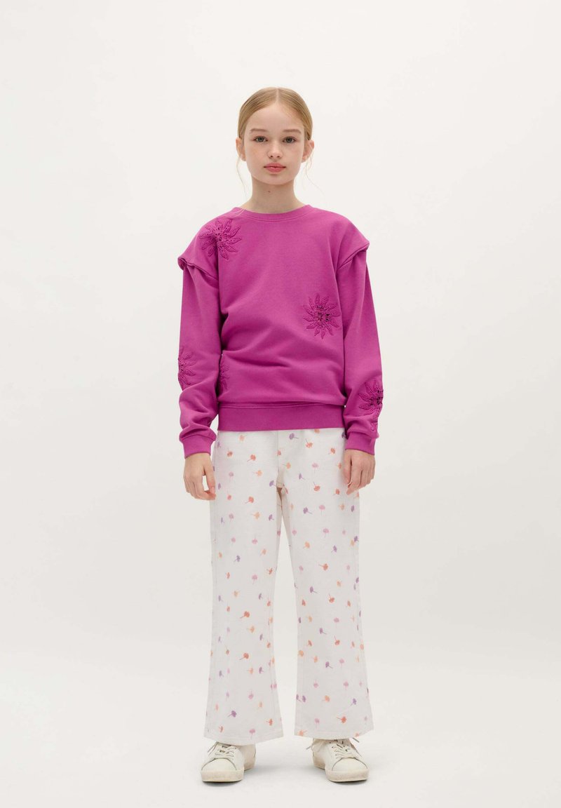 Lilla sweatshirt med blomsterbroderi, parret med hvide bukser med et multicolor blomster mønster. Modellen bærer hvide sneakers.