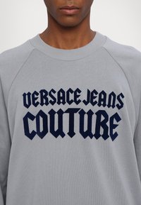 Sivá mikina s námorníckym potlačeným textom "VERSACE JEANS COUTURE" v tučnom, štýlovom písme. Hladký golier a raglánové rukávy.
