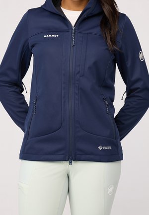 Mujer con una chaqueta softshell azul marino Mammut con capucha y bolsillos con cremallera, combinada con pantalones de color claro.