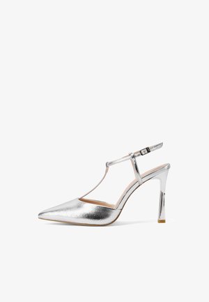 Décolleté argentato metallizzato con punta appuntita, t-bar e fibbia regolabile su sfondo bianco.