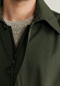 Oliivinvihreä takki, jossa on vetoketjukiinnitys, iso kaulus ja näkyvät napit. Päällä on teksturoitu, kermaisenvärinen neulepaita.