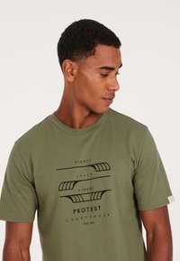 Camiseta de algodón verde oliva con un estampado gráfico negro que presenta el texto "RIGHTY," "LEFTY," y "A-FRAME" en una fuente moderna. Mangas cortas y cuello redondo.