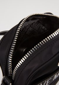 Detailní záběr na otevřenou černou zipovou tašku, která ukazuje stříbrné zipové zuby a černou látkovou podšívku s potiskem.