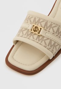 Sandalia tipo chancla beige con dos tiras anchas que presentan un logo MK y un patrón geométrico. Textura suave, punta cuadrada y suela marrón.