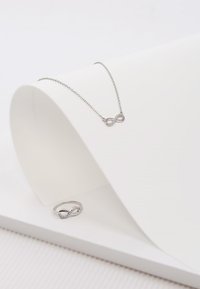 Zilveren infinity-halsketting met kleine kristallen en een bijpassende infinity-ring. Beide stukken hebben een gladde afwerking en liggen op een witte ondergrond.