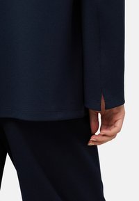 Vêtement bleu marine avec une finition texturée, présentant une fente à l'ourlet et une coupe décontractée. Main légèrement visible, mettant en valeur des ongles naturels.