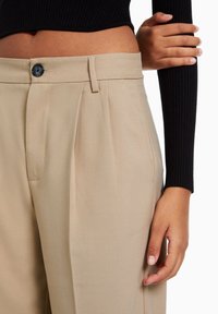 Beige plisserade byxor med en svart knapp vid midjan. Tyget verkar slätt och designen har sidfickor för praktisk användning.