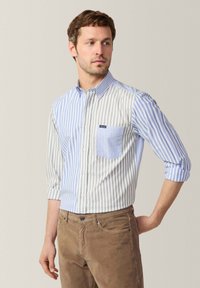 Gestreiftes Baumwoll-Hemd mit Knöpfen, das eine Mischung aus blauen und grauen Streifen aufweist, eine einzelne Brusttasche hat und hochgekrempelte Ärmel hat. Kombiniert mit braunen Hosen.