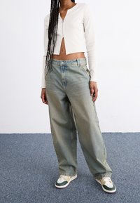 Cardigan corto de canalé ligero, jeans de mezclilla de corte relajado en azul claro y zapatillas con acentos en crema, verde y morado, exhibidos sobre un fondo liso.