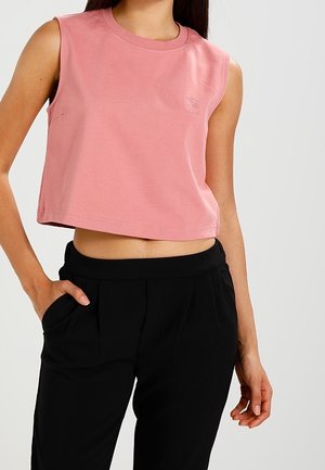 Woman wearing sleeveless pink crop top and high-waisted black pants with one hand in pocket against plain background.