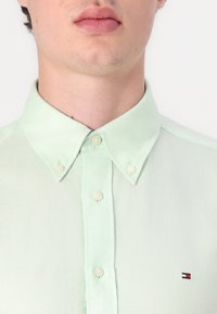 Majica mint zelene boje s dugim rukavima izrađena od lagane tkanine, s oštrim ovratnikom, bisernim gumbima i malim izvezenim logom na prsima.