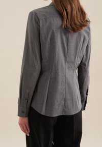 Kvinna med axellångt brunt hår som bär en figurnära grå skjorta med knappar och svarta byxor, visad bakifrån mot en enfärgad bakgrund.