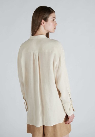 Camicia a maniche lunghe beige chiaro realizzata in tessuto morbido, con colletto alla cinese, piega sul retro e polsini annodati per una vestibilità regolabile.