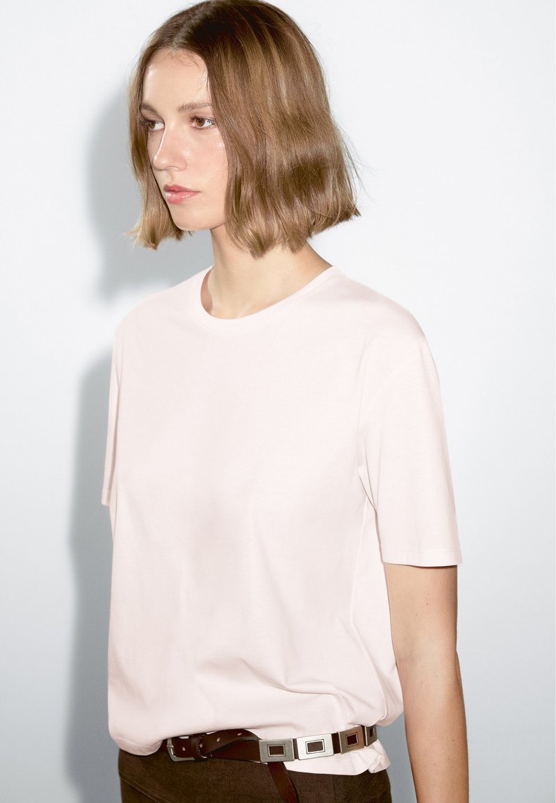 Light pink cotton t-shirt with short sleeves and a round neckline, worn over brown trousers. The shirt appears soft and relaxed in fit.