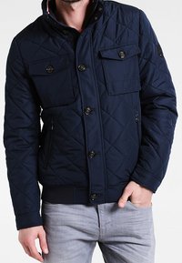 Marinblå quiltad jacka med fyra fickor, knäppning med knappar och ribbade ärmmuddar. Har en hög krage och en subtil struktur i materialet.