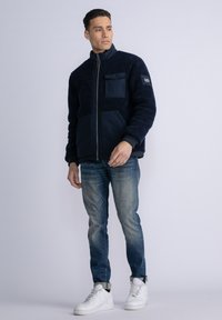 Navy fleece jacket with a textured finish, contrasting front pocket, and high collar. Worn with faded denim and white sneakers.