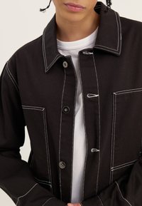 Person som bär en svart jacka med vita söm detaljer över en vit skjorta, med händerna knäppta vid midjan.