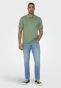 Polo shirt verde a maniche corte abbinato a jeans azzurri chiari e sneaker nere. Il polo presenta un colletto e un design aderente.