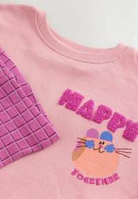 Ružový bavlnený sveter s nadýchaným textom "HAPPY", dizajnom kresleného mačka a detailom rukávu s fialovým kockovaním. Mäkčená textúra so žebrovaným golierom.