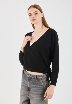Jongeman met een zwarte wikkelsweater over een roze top en versleten grijze spijkerbroek, die de sweater bij de sleutelbeen aanpast tegen een witte achtergrond.