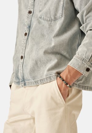 Hellblaues Jeanshemd mit braunen Knöpfen, Brusttasche und hochgekrempelten Ärmeln. Passende cremefarbene Hose und ein Perlenarmband am Handgelenk.