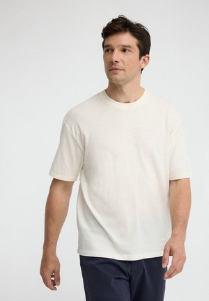 Hombre que lleva una camiseta blanca lisa de manga corta y pantalones oscuros, de pie contra un fondo claro y sencillo, mirando hacia su derecha.
