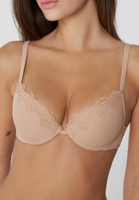 Žena nosící světlobéžovou podprsenku s krajkovým lemem, ukazující horní část trupu, ramena a část obličeje a krku.